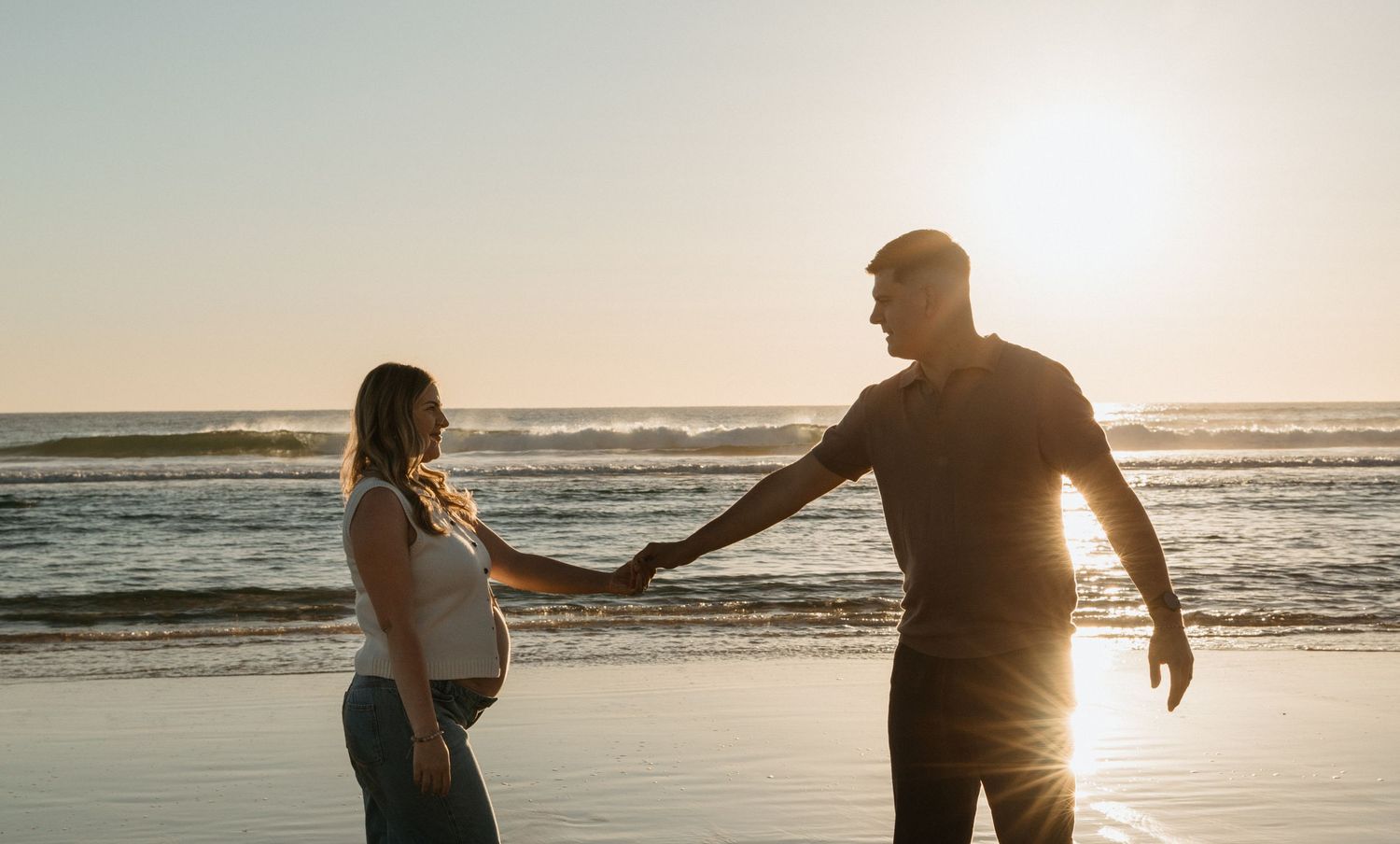 A couple holds hands and walks along a beach at sunset, silhouetted against the golden sky and ocean waves.