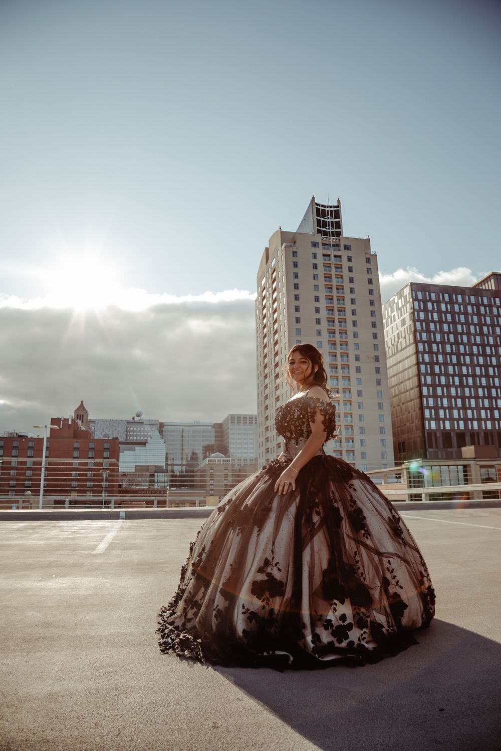 A woman in an elegant brown ball gown poses dramatically against a cityscape backdrop at sunset.