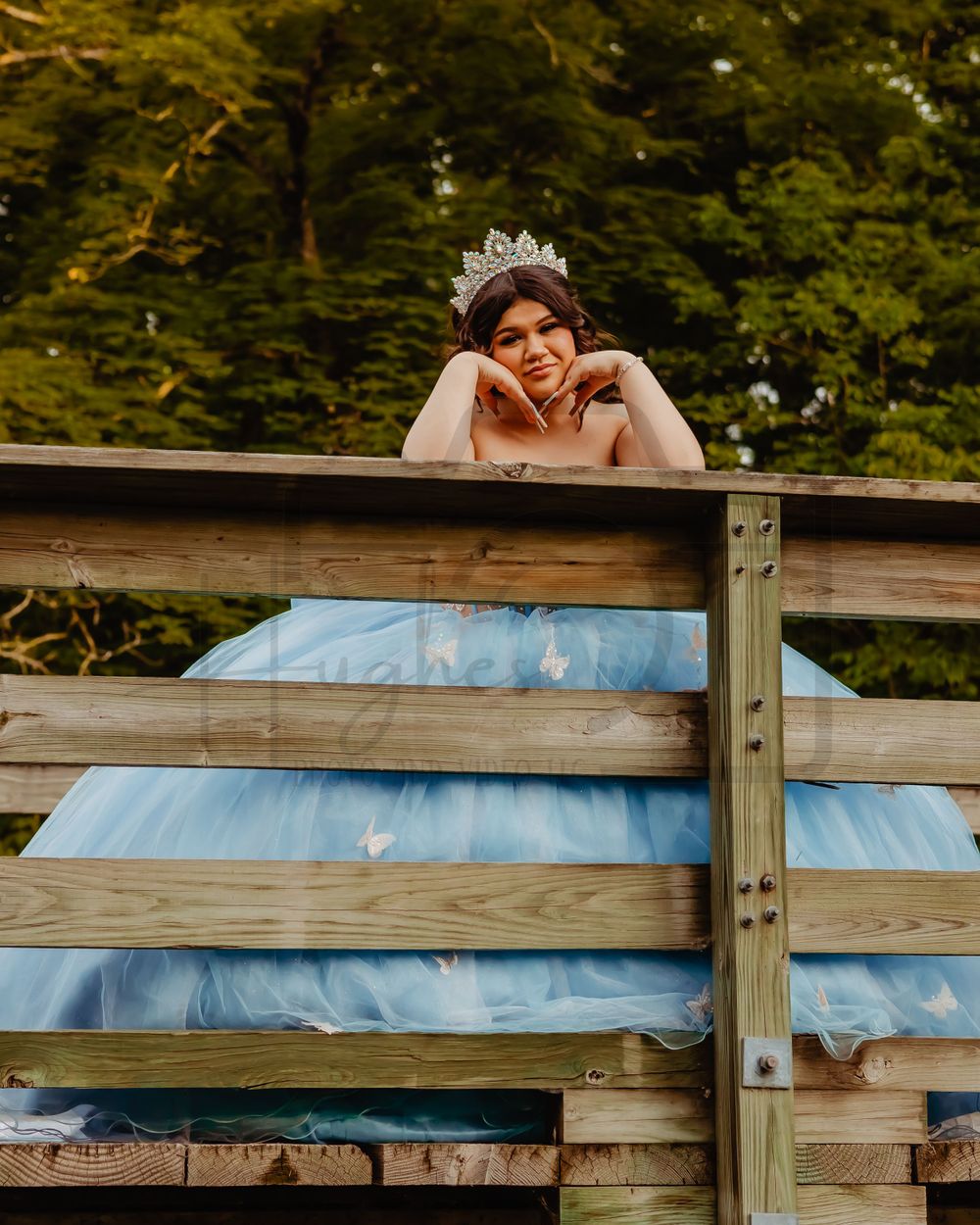 A light blue ballgown and tiara photographed through wooden bridge railings in a forest setting.
