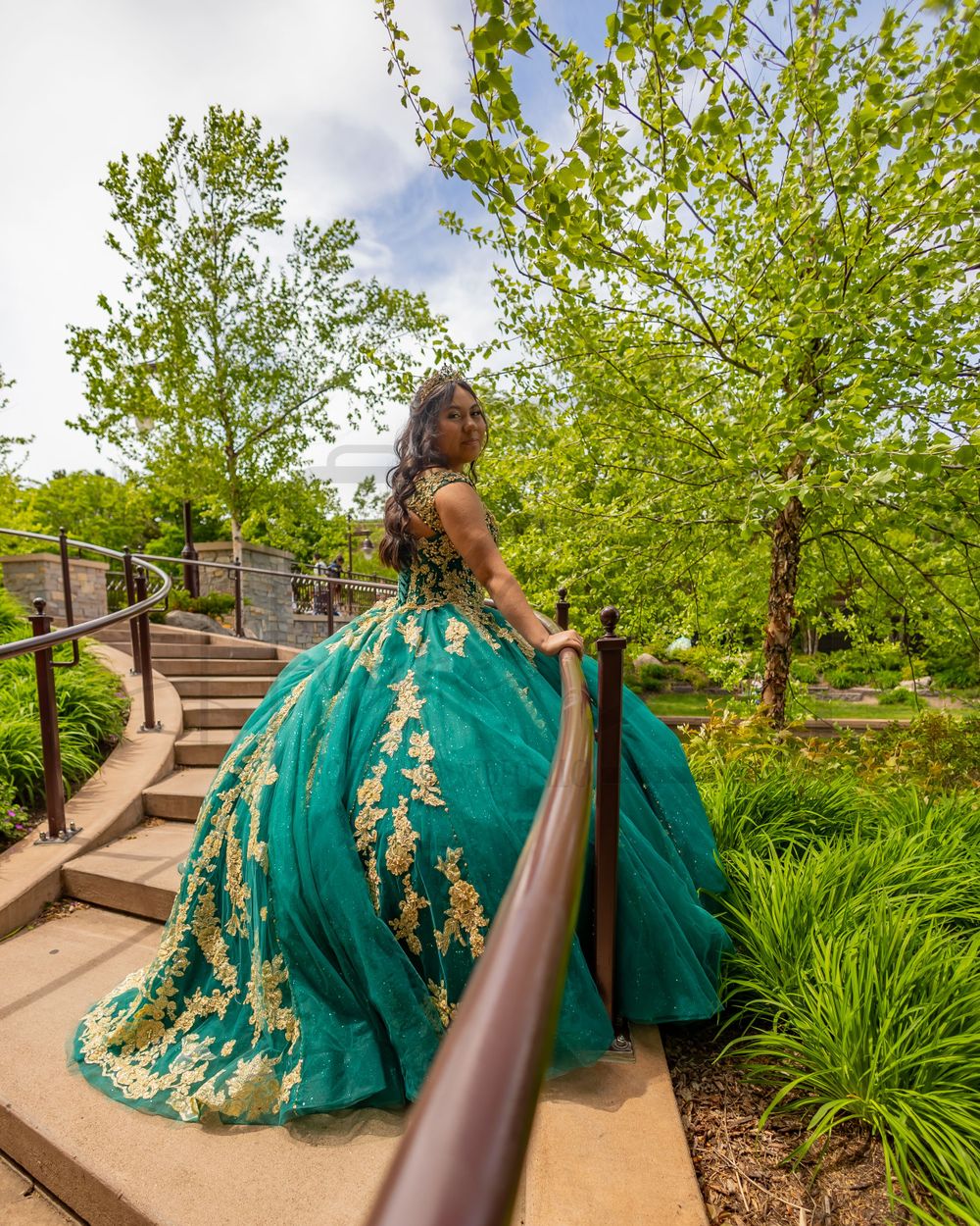 A spiral staircase photo showcasing an elaborate emerald green and gold ballgown in a garden setting.