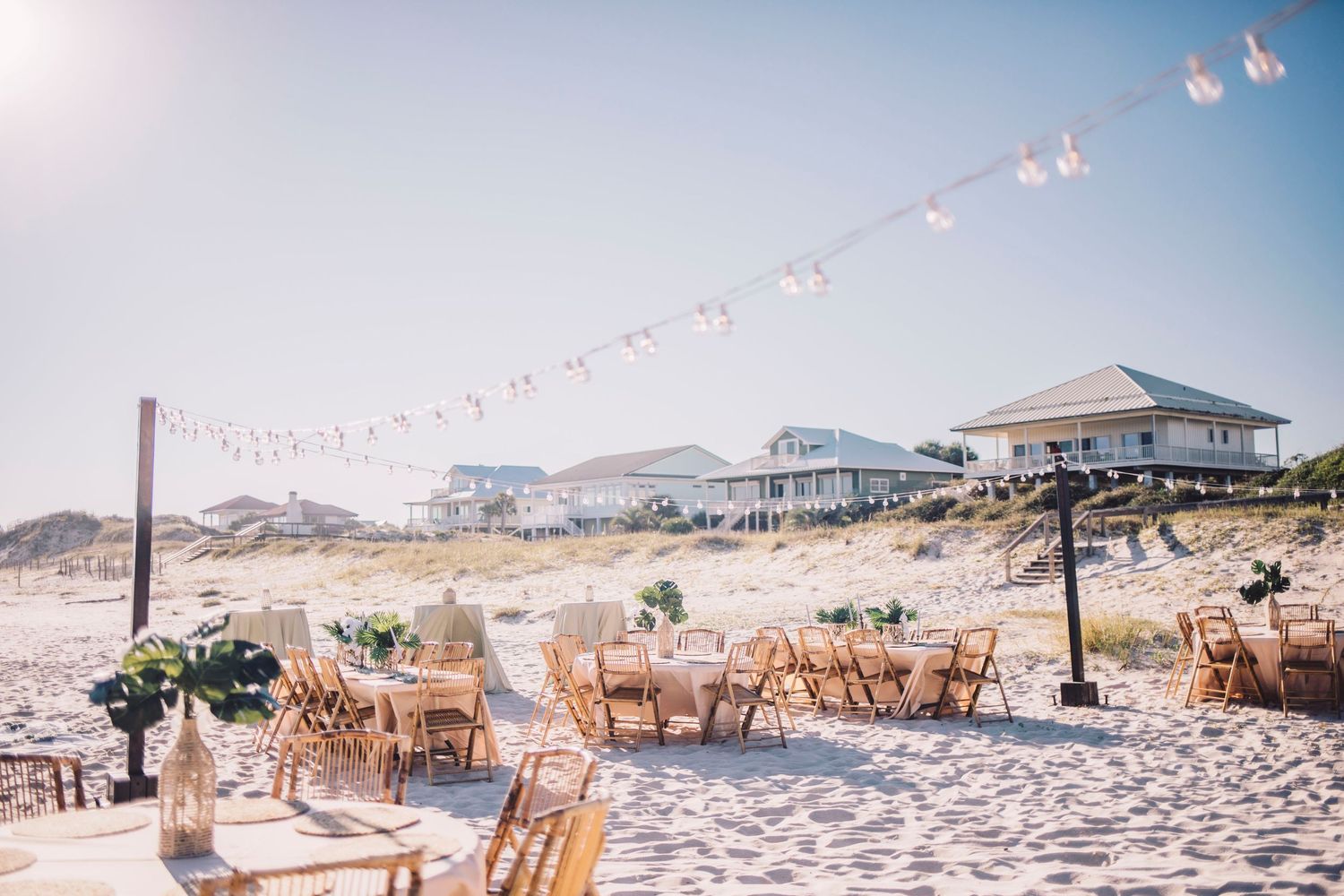 Elegant beach wedding reception setup with wooden chairs, tables, and string lights on sandy shore.