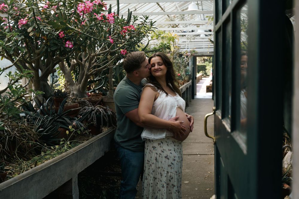 Couple embracing in a lush greenhouse, man kissing woman's cheek among tropical plants and flowers.