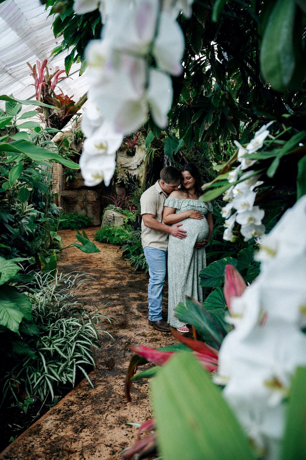 Couple embracing in a lush tropical greenhouse, surrounded by white orchids and green foliage during a maternity photoshoot.