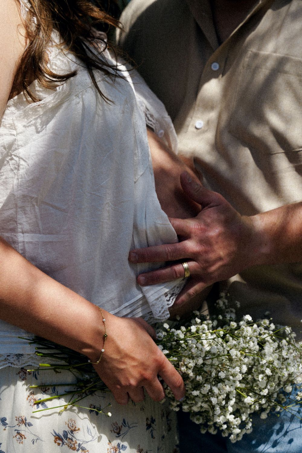 Close-up of couple holding baby bump with white wildflower bouquet during maternity photoshoot.
