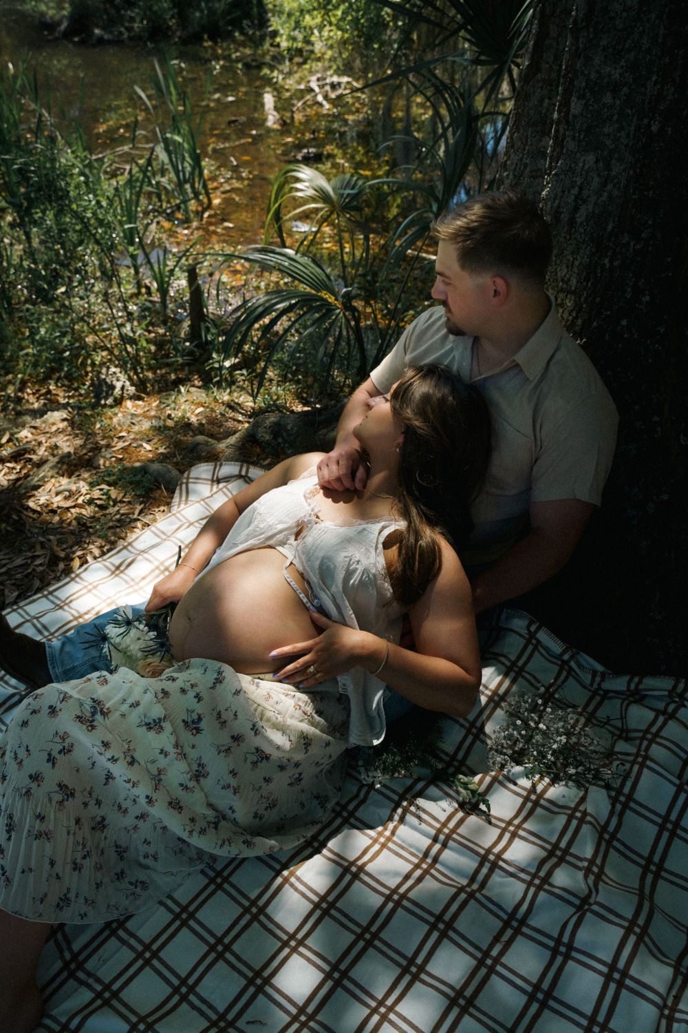 Expecting couple relaxing on picnic blanket by river, woman lying back on partners lap showing baby bump.