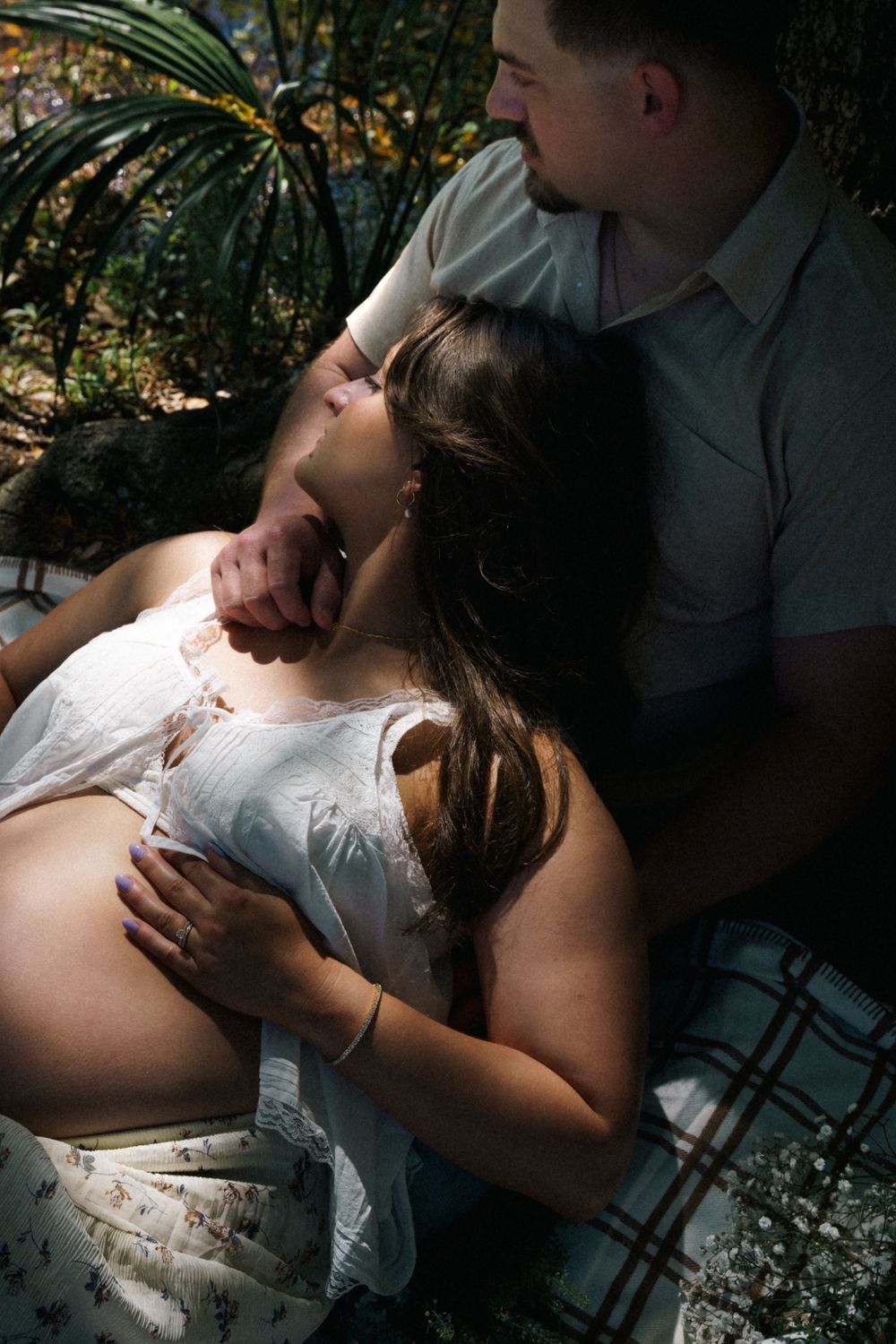 Intimate maternity moment with pregnant woman reclining against partner in dappled sunlight among garden foliage.