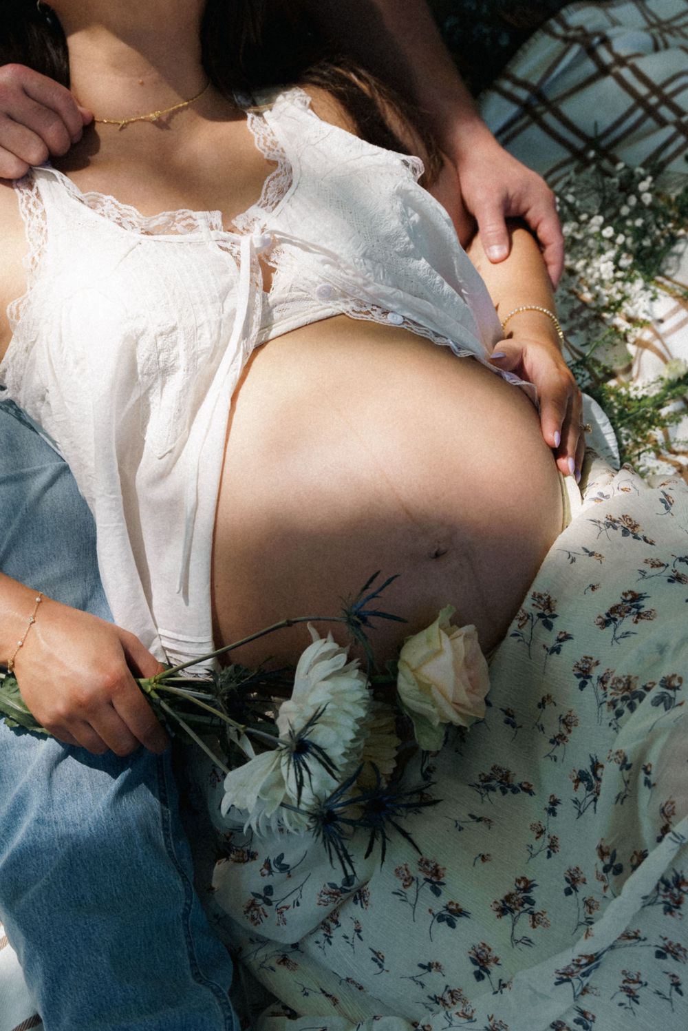 Close-up of pregnant woman's bare belly held by partner's hands, with white roses and floral skirt detail.