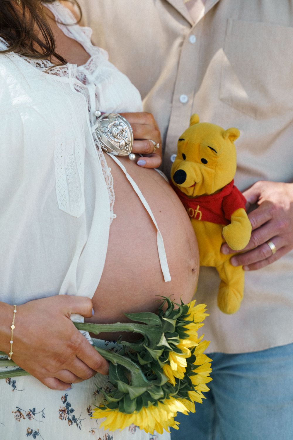 Pregnant woman holding sunflowers with a Winnie the Pooh plush toy placed on her baby bump during maternity shoot.