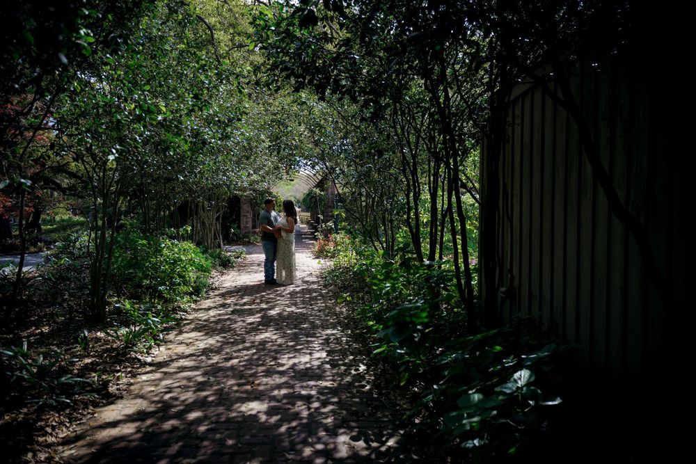 Couple embracing on a shaded garden path surrounded by lush green trees and foliage.