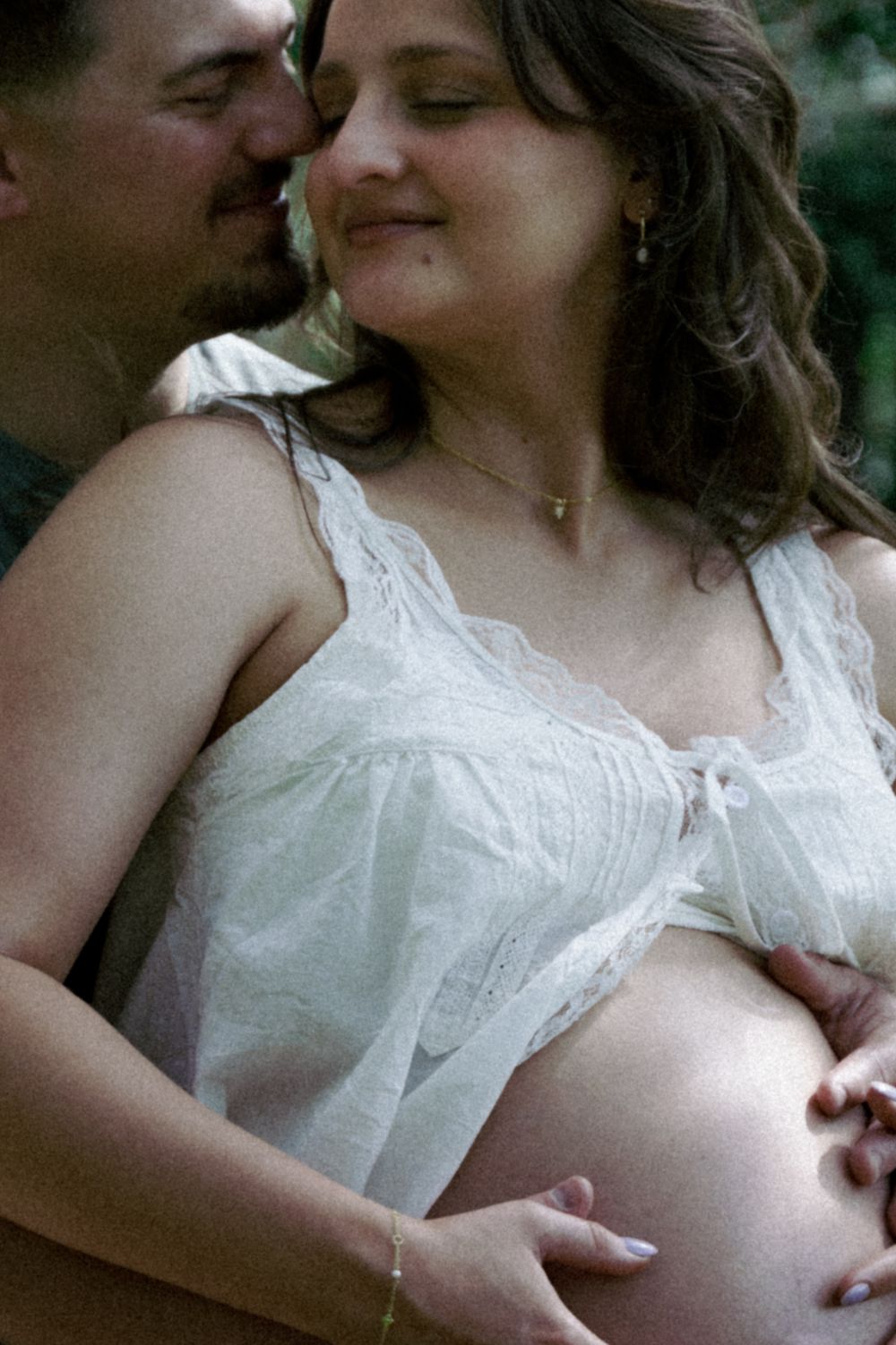 A couple shares a tender moment outdoors, with the man kissing the pregnant woman's cheek as they cradle her bare belly.