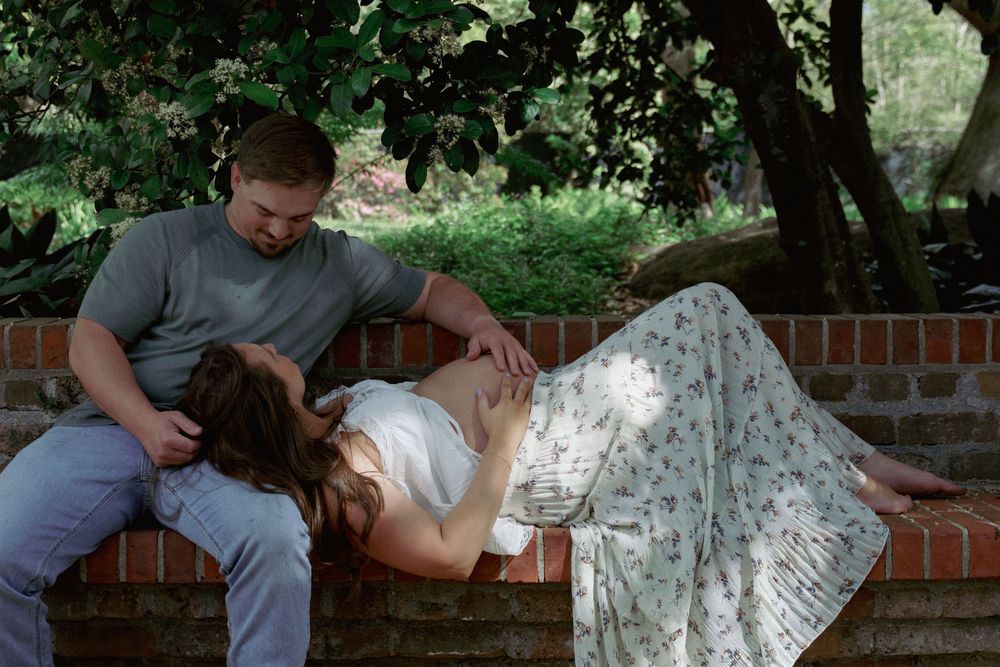 Pregnant woman resting on a brick bench as her partner lovingly touches her baby bump in a garden.