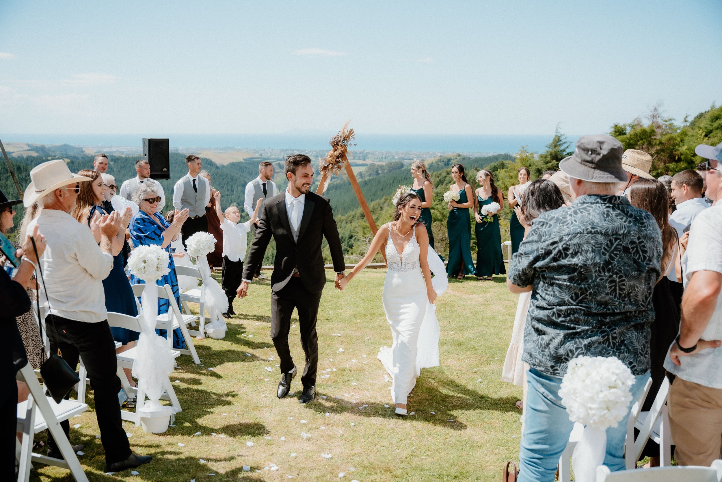 A joyful outdoor wedding ceremony on a hilltop with guests gathered on either side of a grassy path.