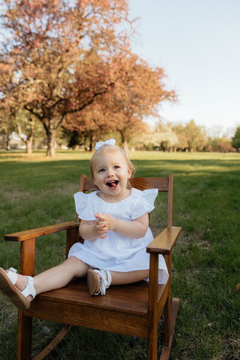 Happy toddler girl in white dress sitting in wooden rocking chair outdoors with autumn trees in background.