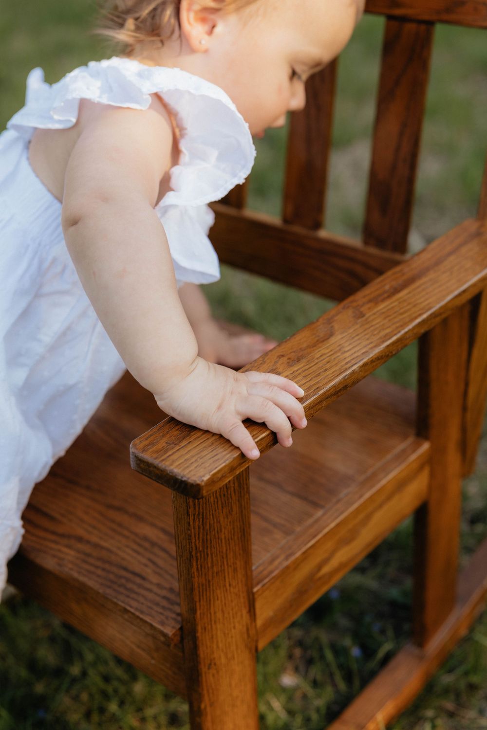A toddler in a white dress grips the arm of a wooden rocking chair outdoors on green grass.