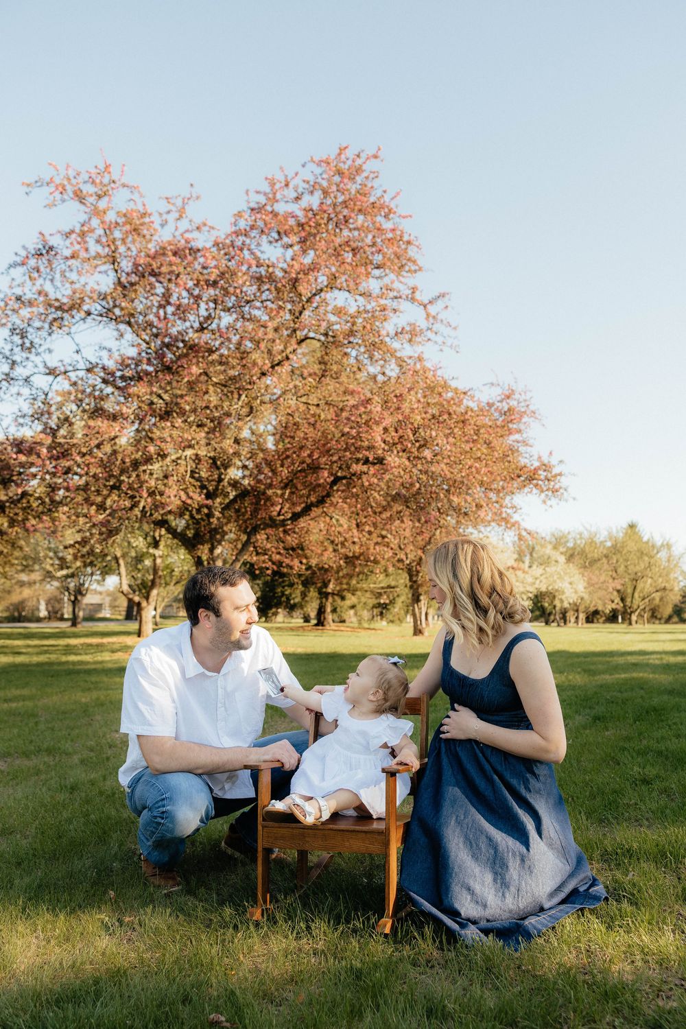 Family with baby in wooden chair under red tree in park.