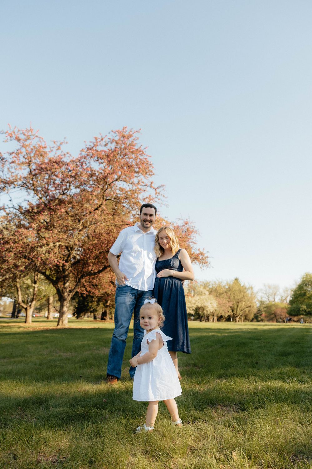 Family with toddler girl standing on grass near red tree outdoors.