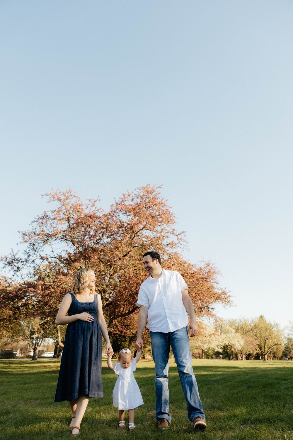 A family of three walks in a park, parents holding toddler's hands, with autumn trees and blue sky behind them.