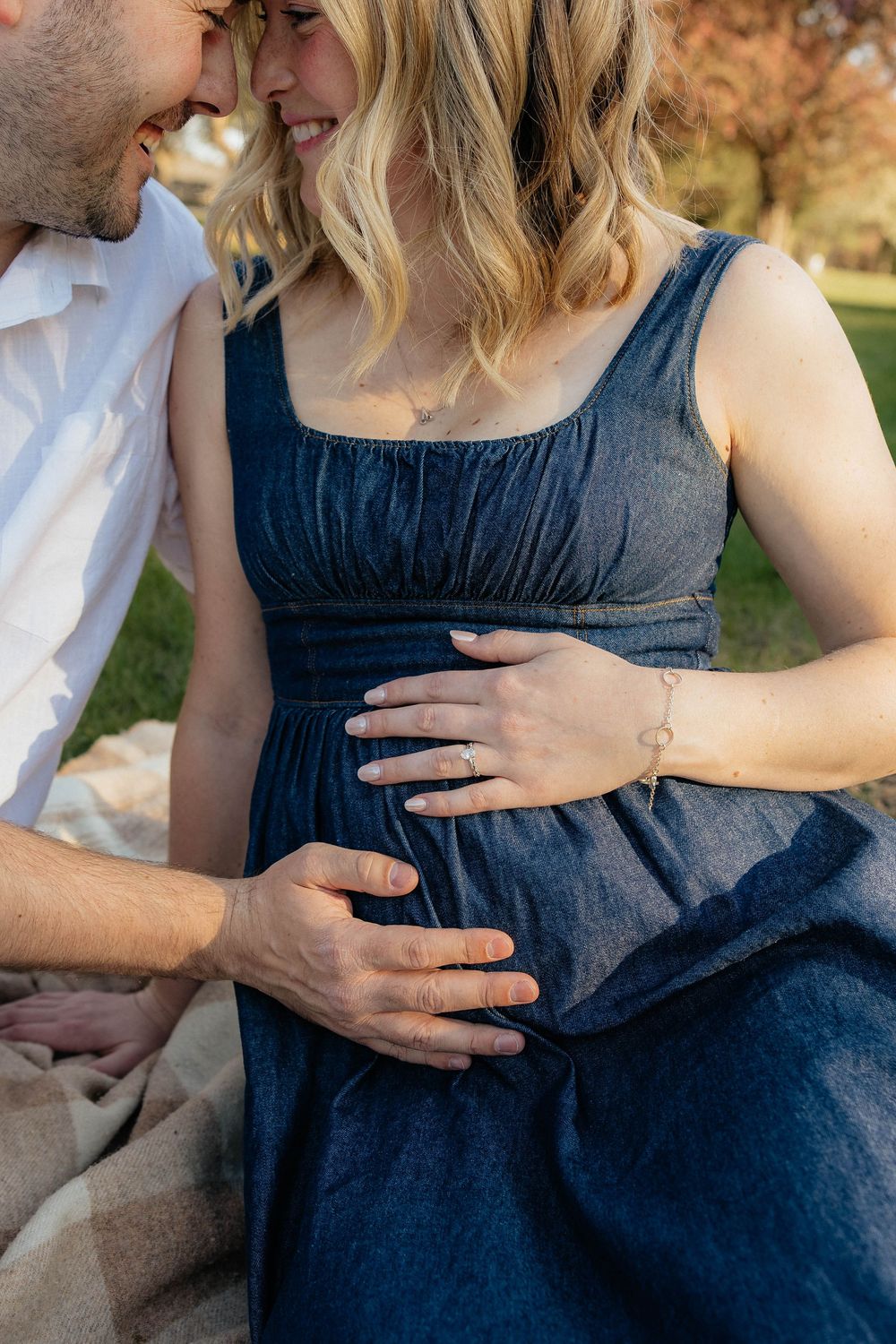 A couple tenderly cradles a pregnant belly outdoors, with the woman wearing a navy dress and a delicate bracelet.