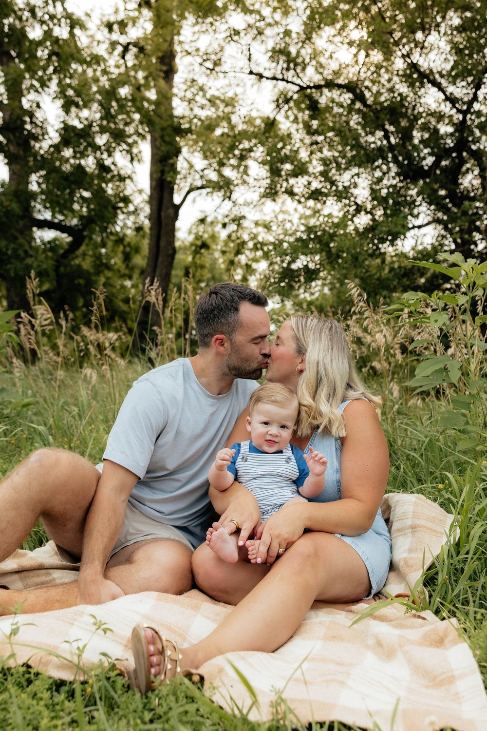 Family relaxing together on picnic blanket in grassy field with trees and sunlight in background.