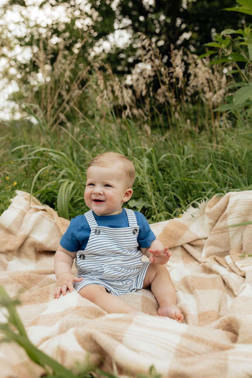 A happy baby wearing blue striped overalls sits on a beige blanket outdoors surrounded by greenery.