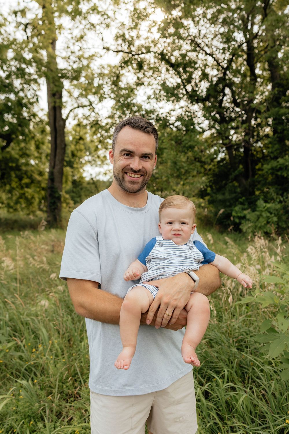 A happy adult and baby stand together in a sunlit grassy field surrounded by trees on a summer day.