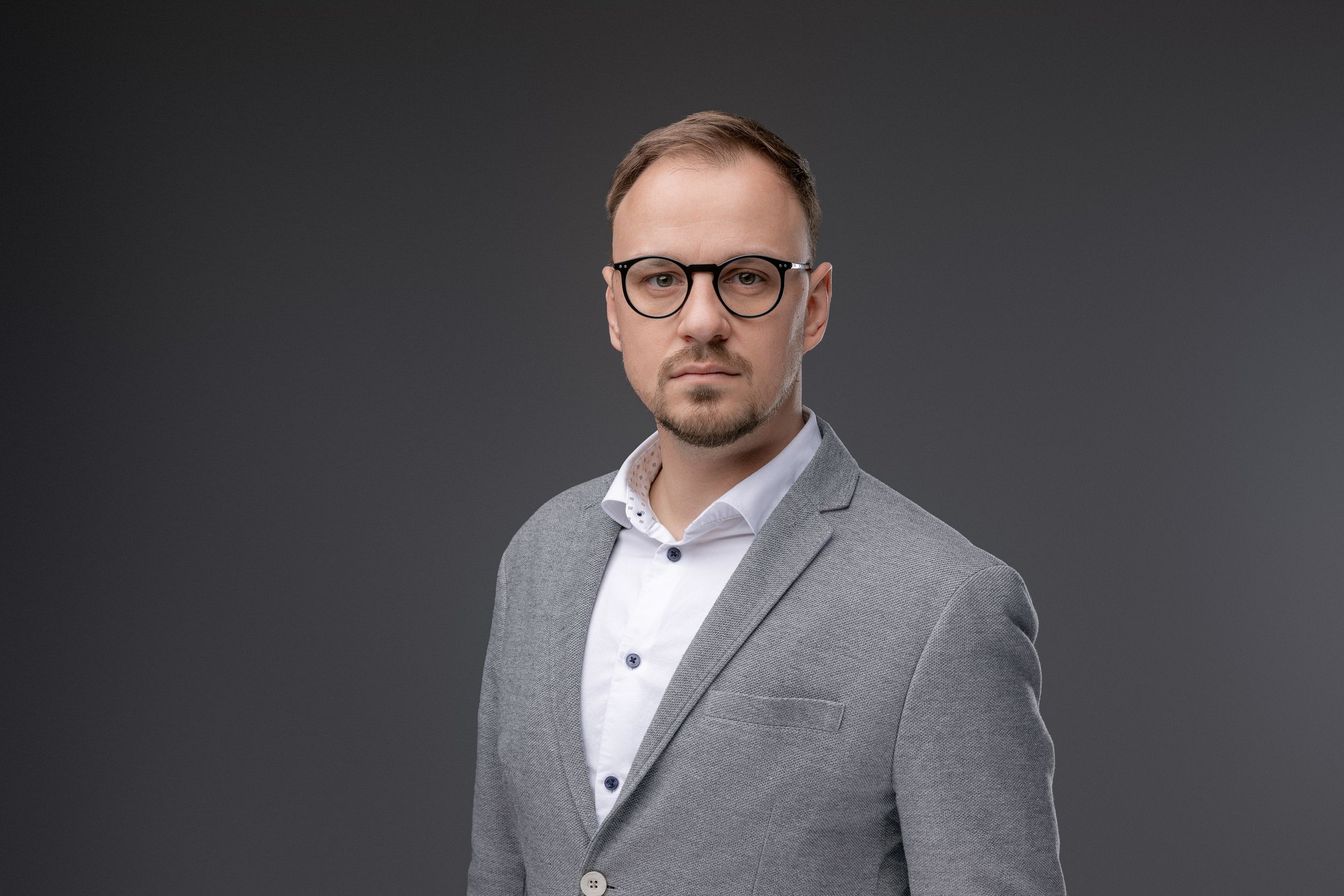Professional headshot of businessman wearing grey suit jacket and glasses against dark background.