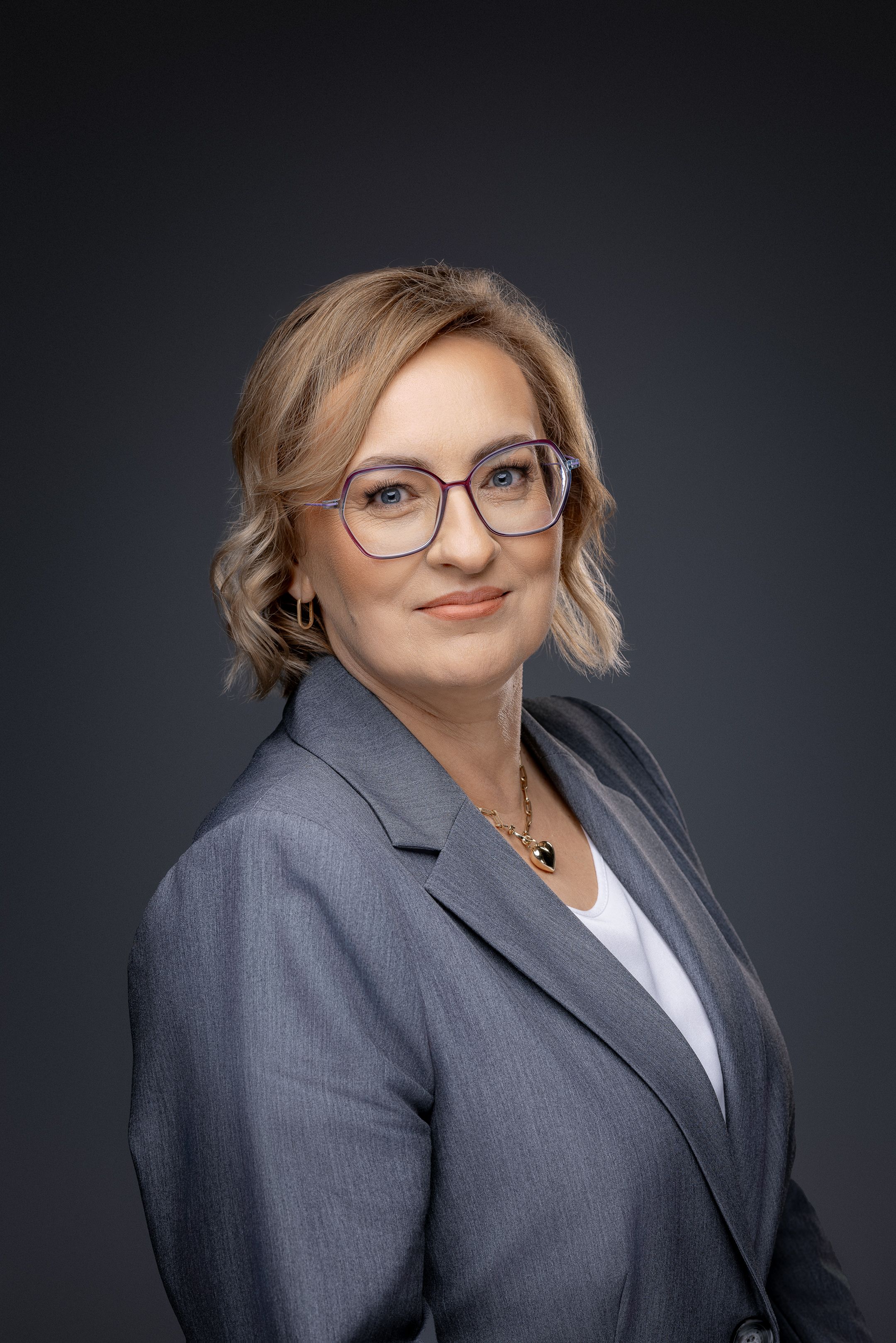 Professional headshot of businesswoman in grey blazer and glasses against dark background.