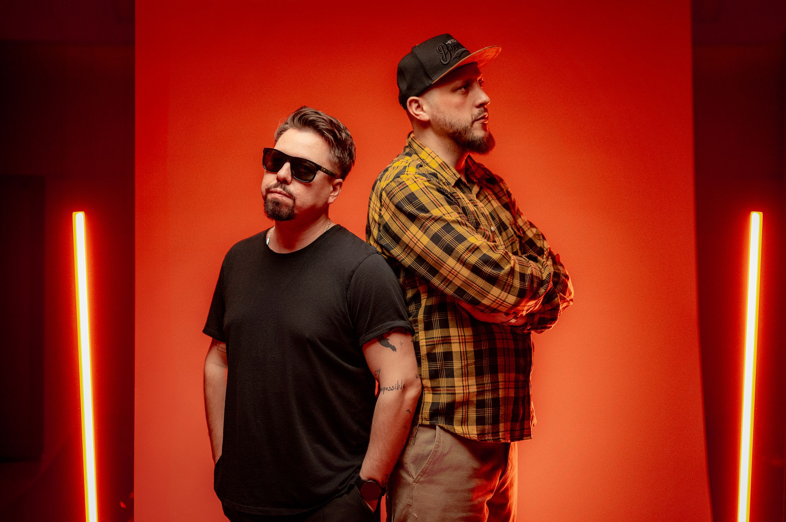 Two musicians posed back-to-back against red background with vertical light bars.