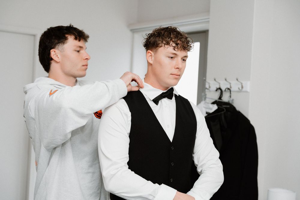 Two formally dressed people getting ready in a room with one adjusting the other's outfit.