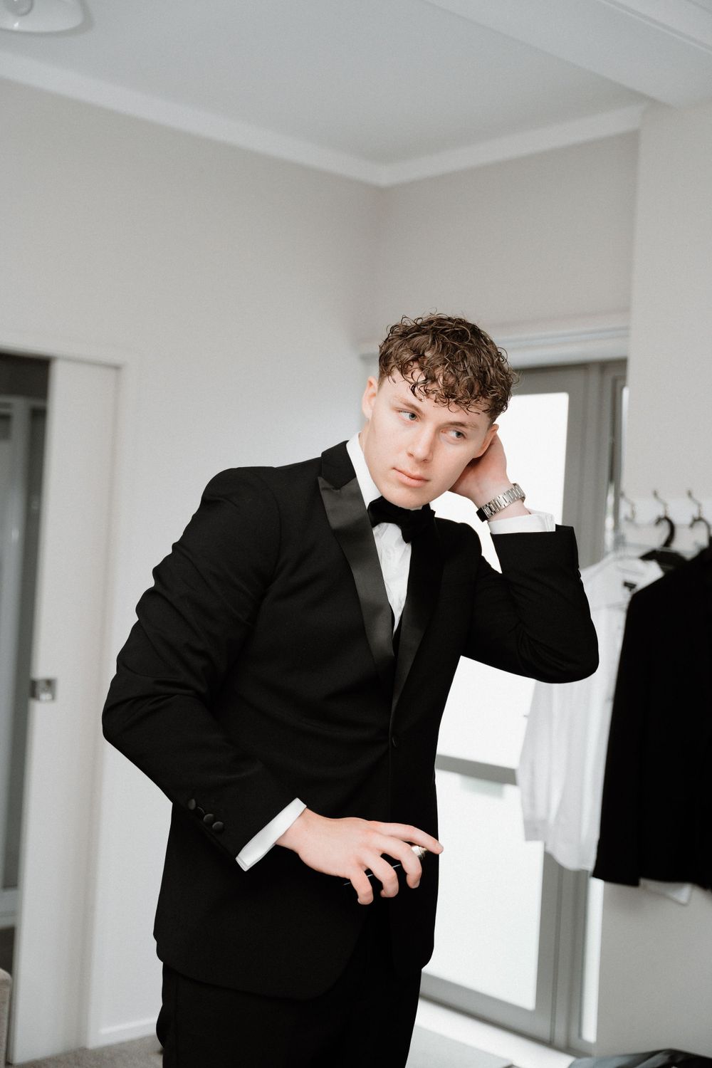 Person in black tuxedo and bow tie poses pensively in a dressing room.