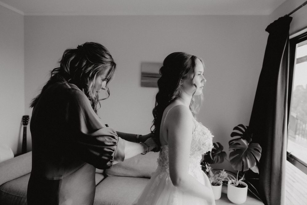 A candid black and white photo showing final wedding dress preparations in a sunlit room.