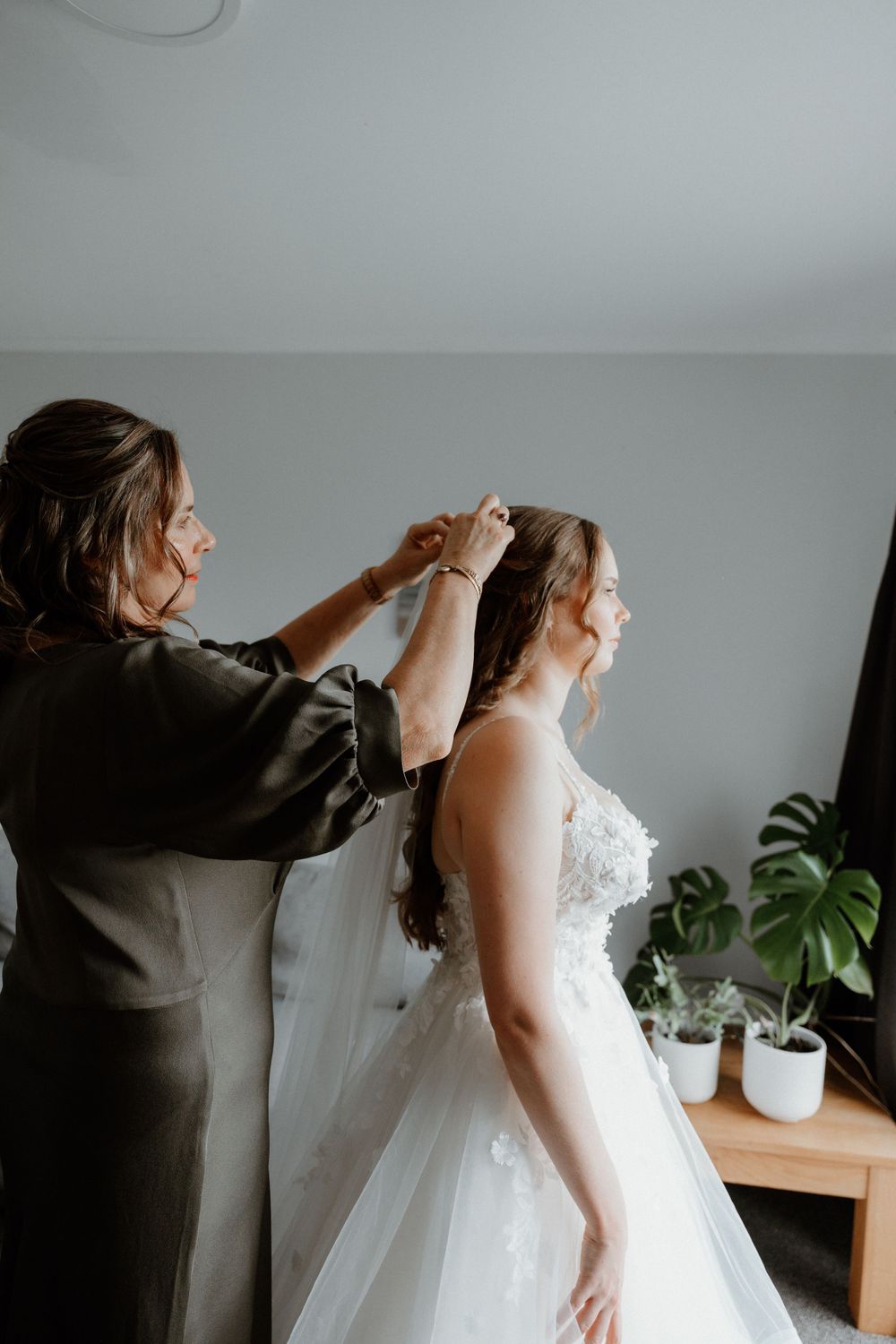 Bride in white lace wedding dress gets hair styled in front of potted monstera plant during wedding preparations.