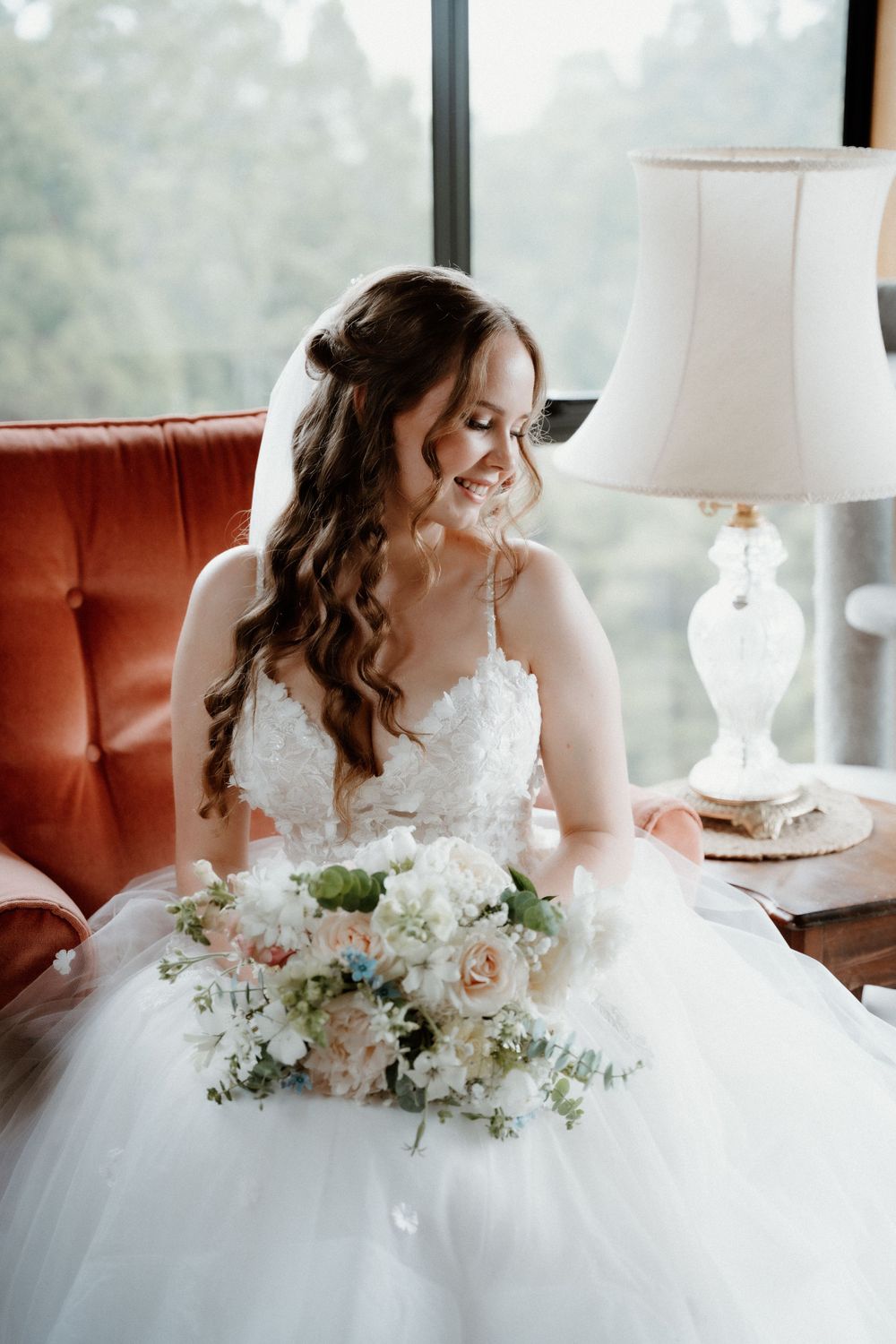 A bride in white tulle wedding gown sits on orange chair holding delicate bouquet of blush and white flowers.