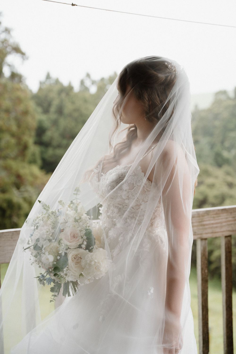 A bride in a flowing white veil and dress holds a delicate bouquet of white flowers against a natural outdoor backdrop.