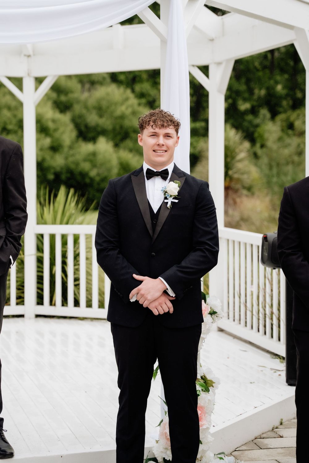 A groom in a classic black tuxedo and bowtie smiles while standing under a white outdoor wedding gazebo.