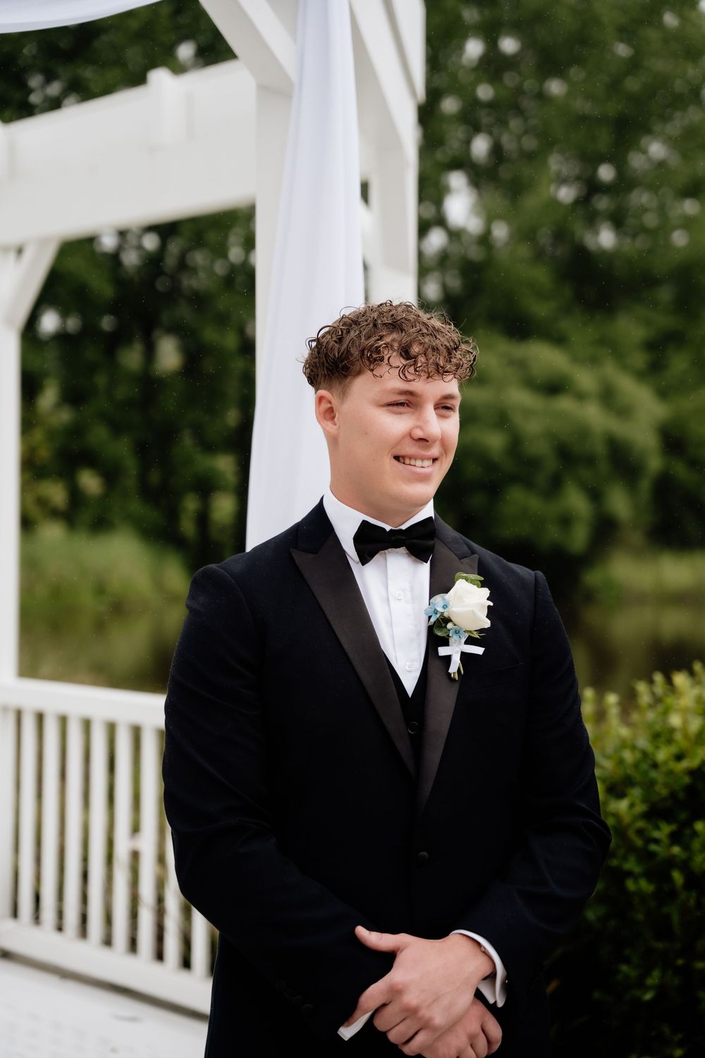 Wedding portrait of a groom in a black tuxedo and bow tie with boutonniere standing by a white gazebo.
