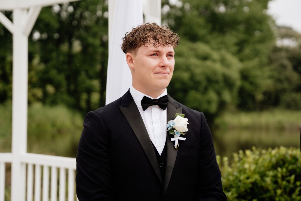 A groom in a black tuxedo and bow tie stands outdoors near a white arbor with greenery in the background.