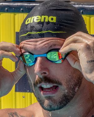 Competitive swimmer adjusting mirrored goggles and wearing black Arena swim cap during race preparation.