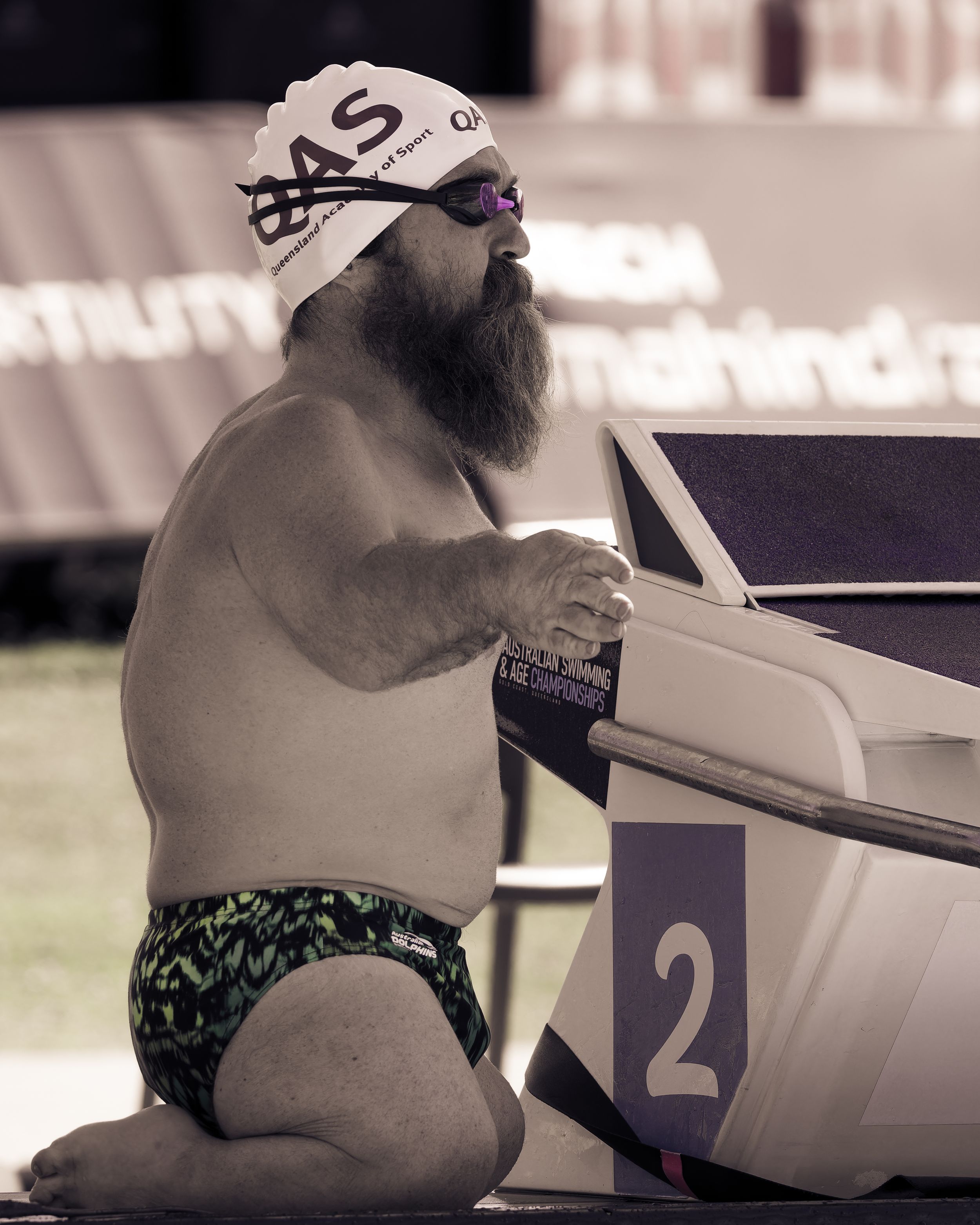 Bearded swimmer in white cap prepares at starting block number 2 during competition.