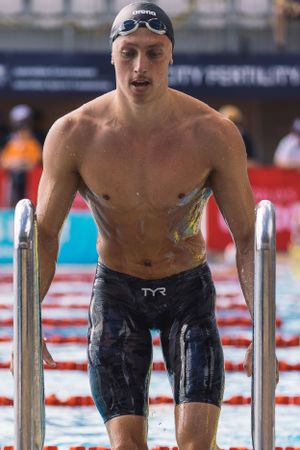 Athlete in black jammers and swimming cap exits pool using metal handrails.