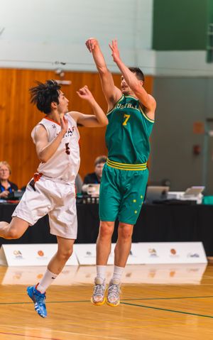 Two basketball players jumping during game action, one shooting in green jersey and one defending in white.