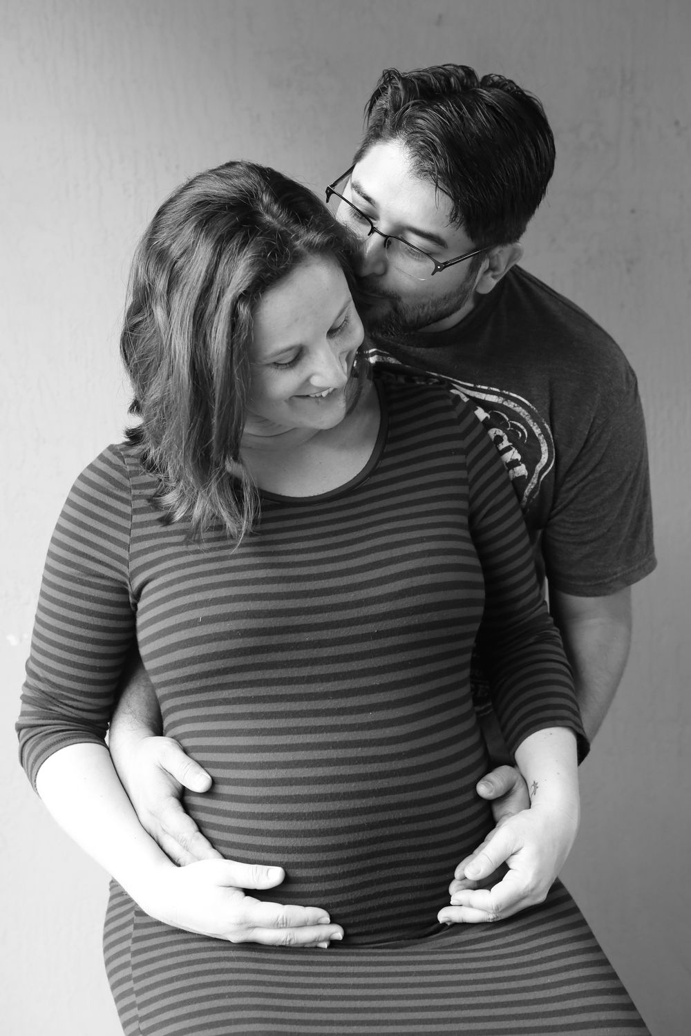 Black and white maternity portrait showing couple embracing, wearing casual striped clothing.