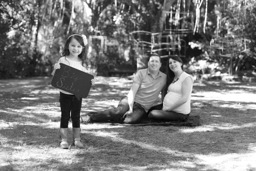 Outdoor family maternity photo with child holding sign in wooded area.