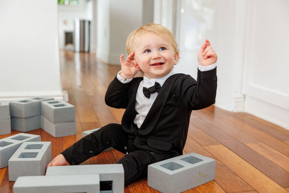 Adorable toddler in a formal black tuxedo plays with building blocks on hardwood floor.