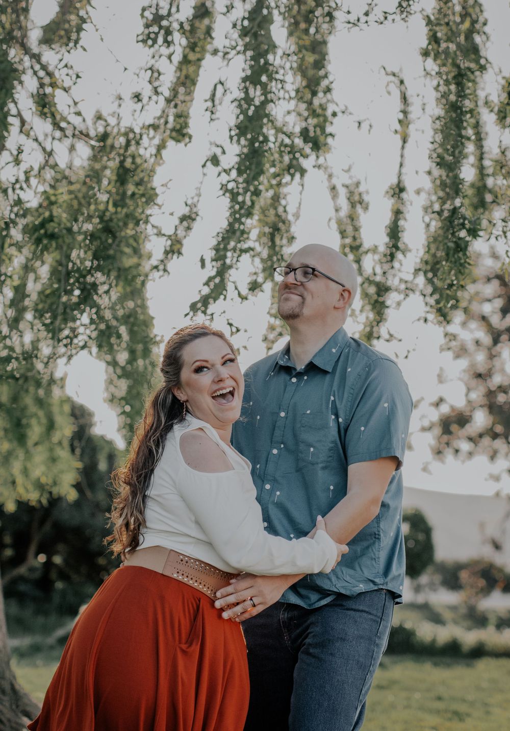 Joyful engagement photo of couple laughing under weeping willow tree.