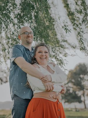 A joyful couple embraces under a willow tree in casual outdoor attire during a sunset photoshoot.