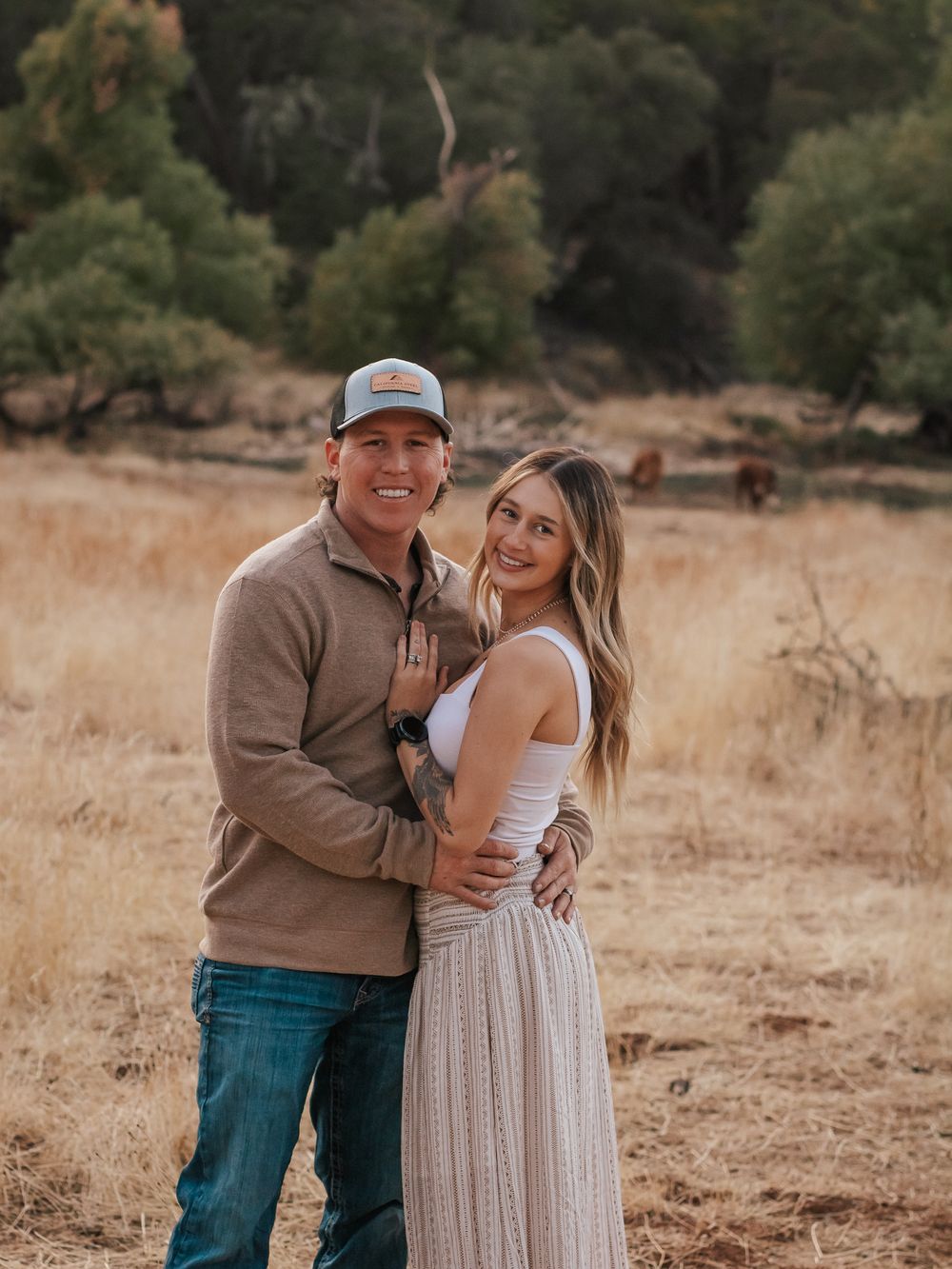 A couple poses together in a rustic field setting wearing casual fall attire during golden hour.