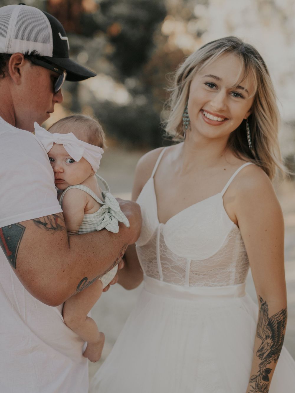 A couple in white attire poses with their baby during an outdoor photoshoot.