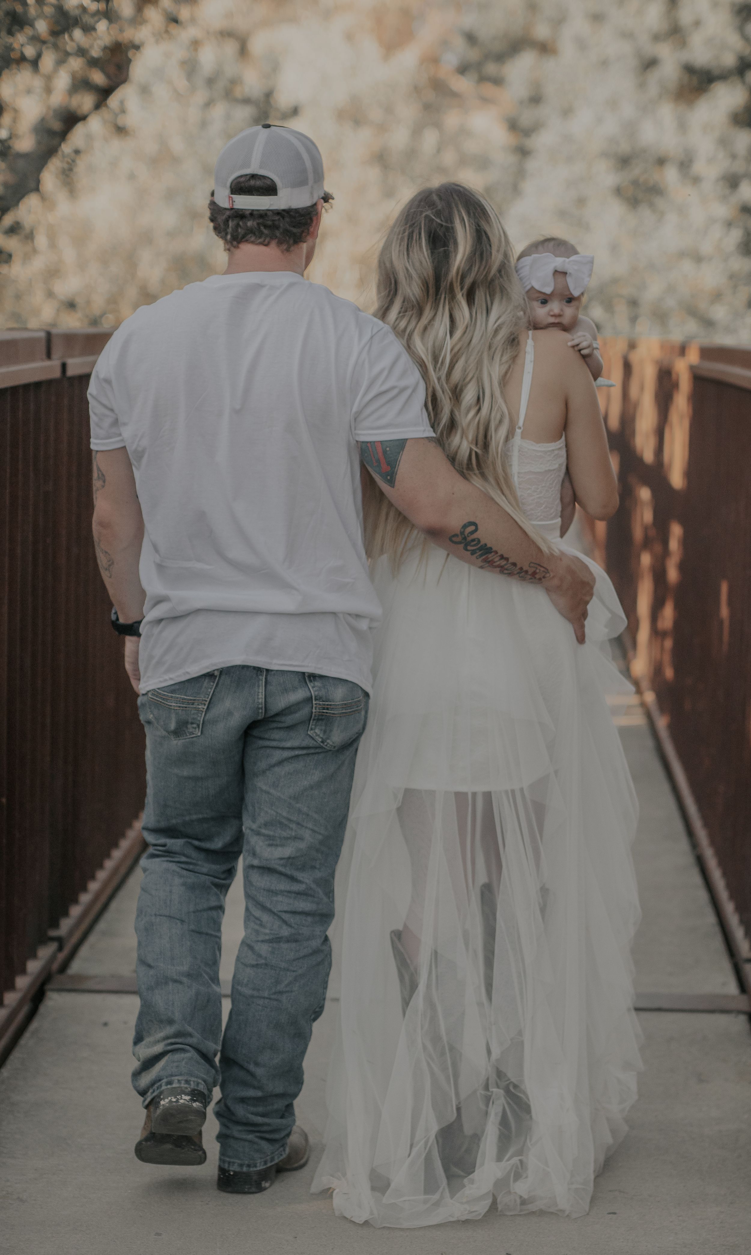A couple walking on a bridge together with the person in a flowing white dress and baseball cap.