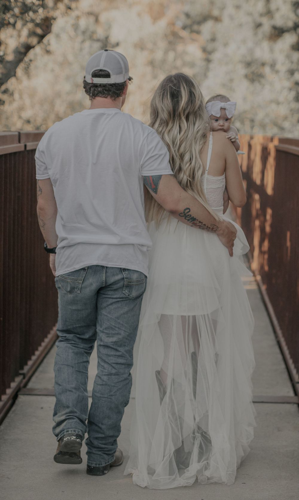 A couple walking on a bridge together with the person in a flowing white dress and baseball cap.